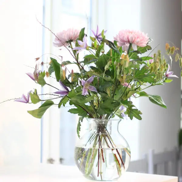 Lockerer Strauß mit rosa Dahlien und lila Clematis in einer klaren, runden Glasvase am hellen Fenster. 4