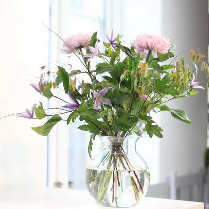 Lockerer Strauß mit rosa Dahlien und lila Clematis in einer klaren, runden Glasvase am hellen Fenster. 4