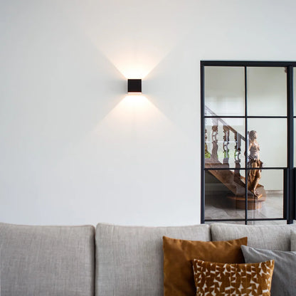 Schwarze Würfel-Wandlampe an weißer Wand über einem grauen Sofa mit Kissen, daneben eine Glastür mit Stahlrahmen. 10
