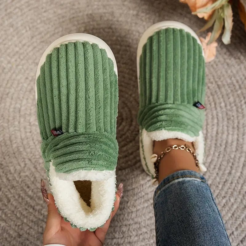 Grüne gerippte Hausschuhe von oben, ein Paar an einer Hand gehalten, der andere Schuh am Fuß in Jeans.