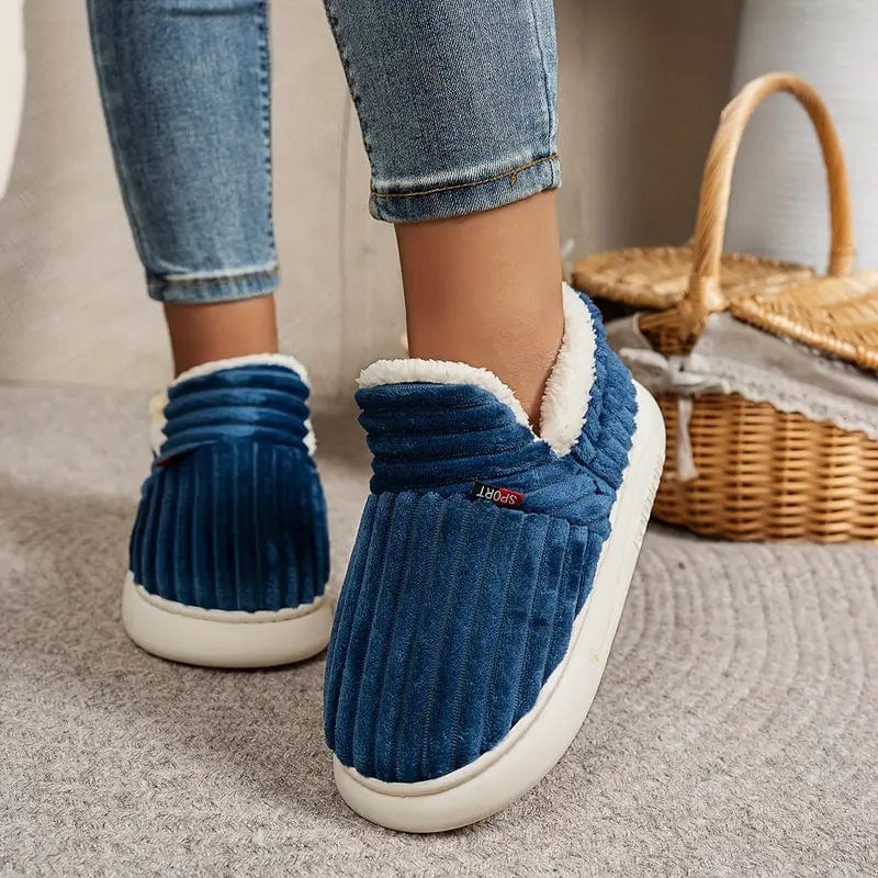 Blaue gerippte Hausschuhe an den Füßen einer Person in Jeans, daneben ein Weidenkorb auf grauem Teppich.