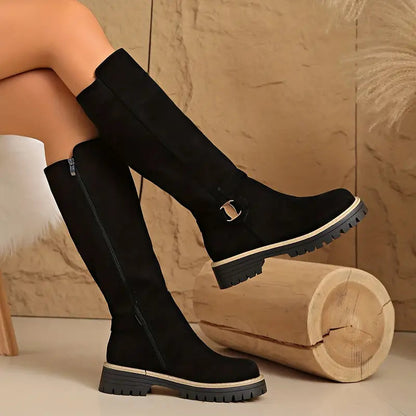 Schwarze kniehohe Stiefel mit seitlichem Reißverschluss und goldfarbener Schnalle, an überkreuzten Beinen.