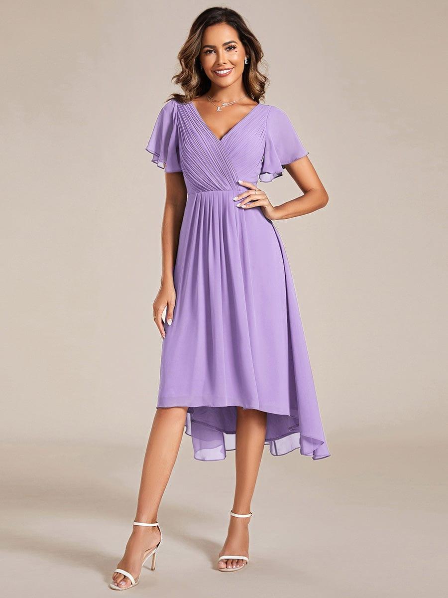 Model in fliedervioletten Chiffonkleid mit plissiertem V-Ausschnitt, Flatterärmeln und Hoch-Tief-Saum, Hand an der HĂĽfte, weiĂźe Sandaletten. 100