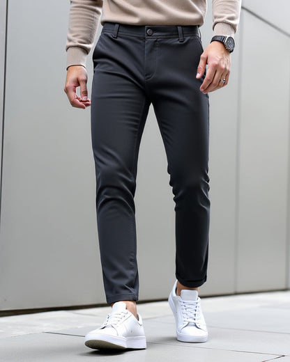 Dunkelgraue Chino vor grauer Wand mit beigefarbenem Strickpullover und weißen Sneakern, Beinabschluss leicht umgeschlagen.