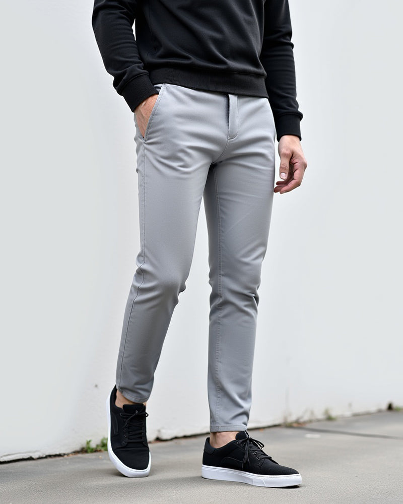 Hellgraue Chino in einer weiteren Ansicht im Freien mit schwarzem Pullover und schwarzen Sneakern mit weißer Sohle.
