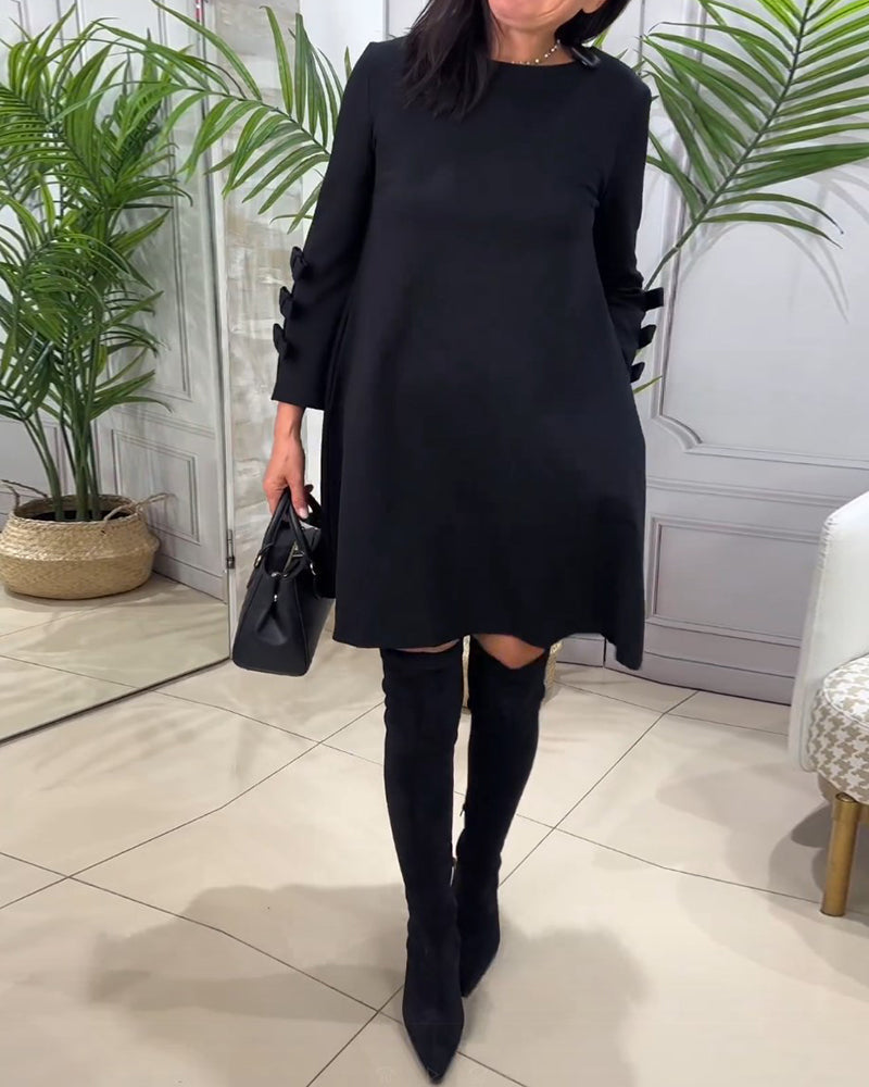 Frau in schwarzem Kleid und Stiefeln, hält schwarze Handtasche, steht in modernem Raum.