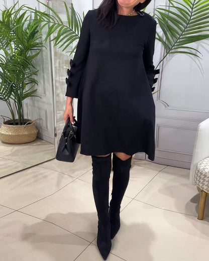 Frau in schwarzem Kleid und Stiefeln, hält schwarze Handtasche, steht in modernem Raum.
