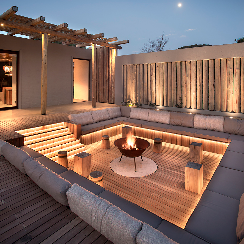 LED-beleuchtete Terrasse mit Feuerstelle, Holzsitzbereich und moderner Pergola bei Abenddämmerung.