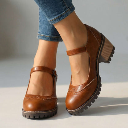 Braune Mary Janes mit verstellbarem Riemen, Lochmuster und profilierter Sohle, zu Jeans getragen, vor neutralem Hintergrund. 2
