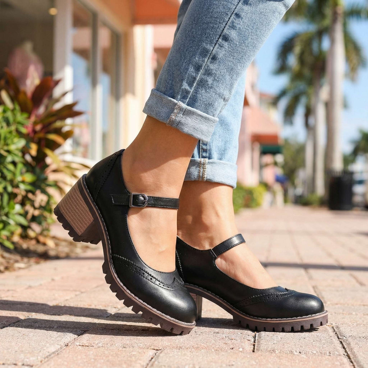 Schwarze Mary Janes mit Schnalle zu hochgekrempelten Jeans, getragen auf einem gepflasterten Gehweg zwischen Palmen und Pflanzen. 4