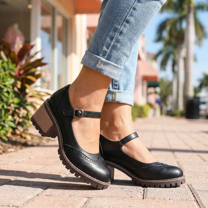 Schwarze Mary Janes mit Schnalle zu hochgekrempelten Jeans, getragen auf einem gepflasterten Gehweg zwischen Palmen und Pflanzen. 4