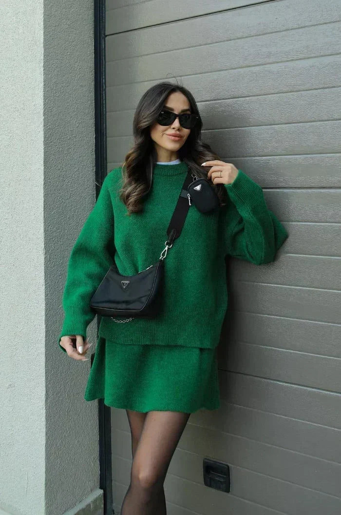 Model lehnt in grünem Pullover und Rock an einer Wand, eine Hand am Kragen, schwarze Umhängetasche über der Schulter. 10