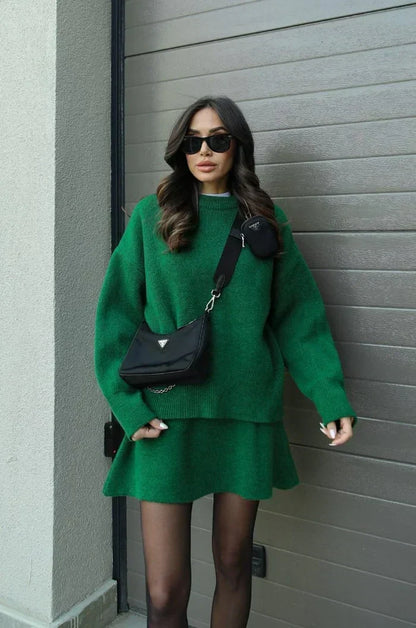 Model in grünem Strick-Set aus Pullover und ausgestelltem Minirock, mit schwarzer Umhängetasche vor einem Garagentor. 8