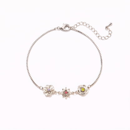 Silberfarbenes Armband mit drei Blumenanhängern und violettem, rotem und grünem Stein, flach auf hellem Hintergrund. 16