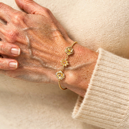 Nahaufnahme einer reiferen Hand mit dem goldenen Blumenarmband, der Ärmel eines cremefarbenen Pullovers ist zu sehen. 11