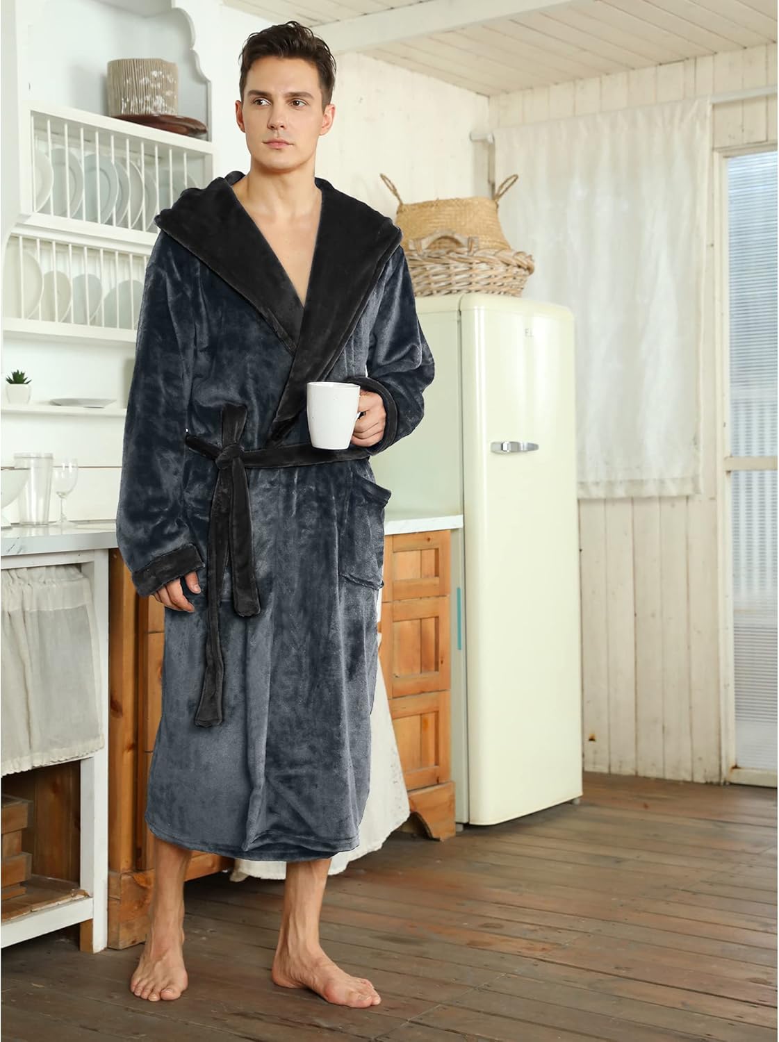 Mann im grauen Fleece-Bademantel mit Kapuze, hält Tasse, steht in rustikaler Küche.
