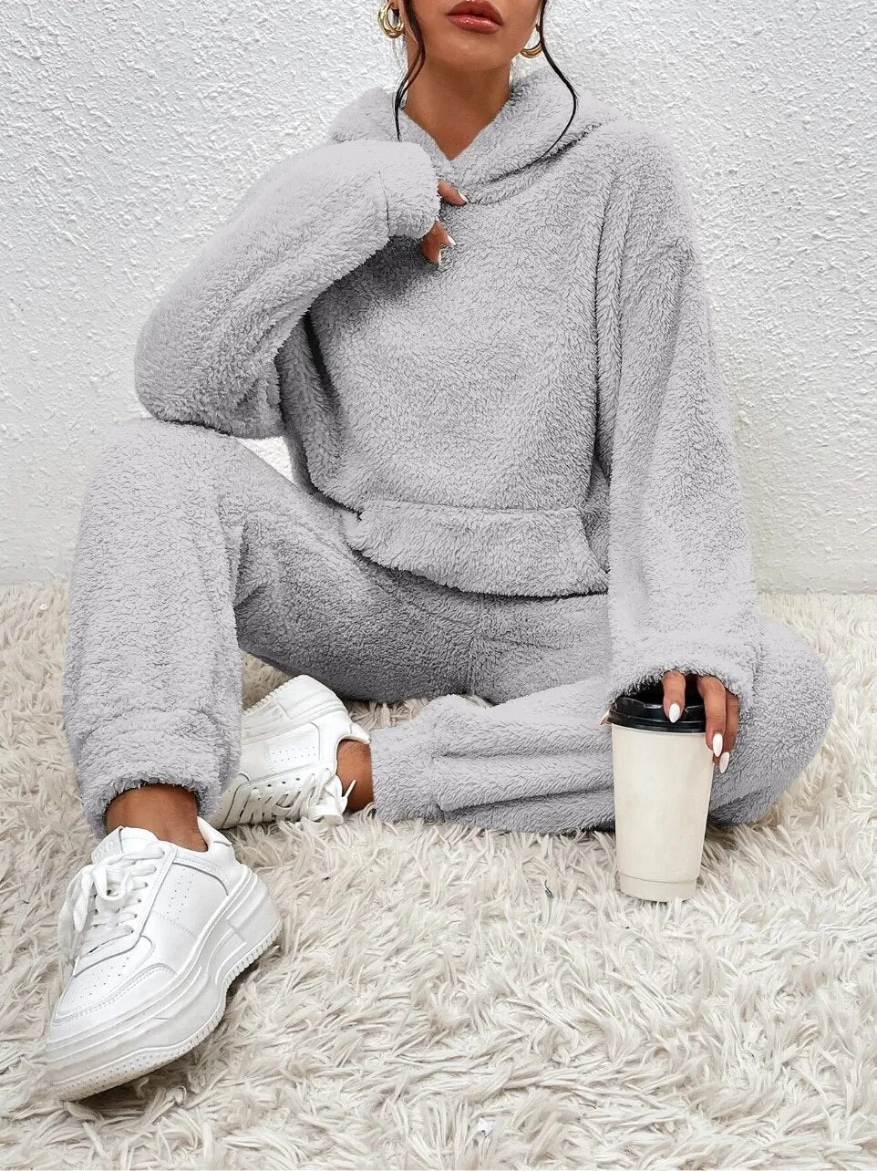 Frau in grauem Fleece-Trainingsanzug, weiße Sneaker, hält Kaffeebecher, sitzt auf Teppich.