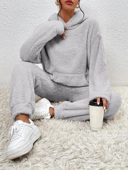 Frau in grauem Fleece-Trainingsanzug, weiße Sneaker, hält Kaffeebecher, sitzt auf Teppich.