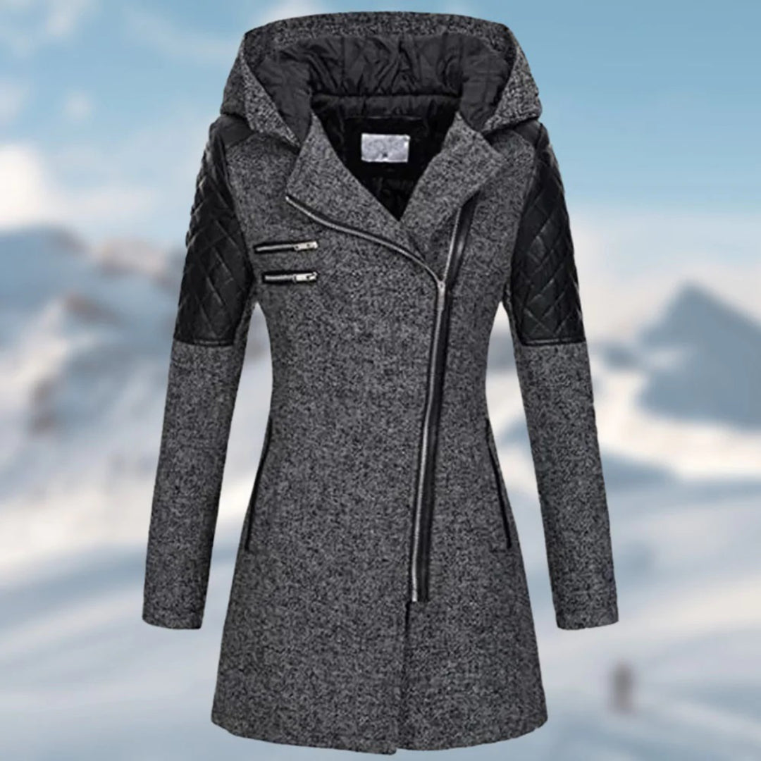 Grauer Damen-Wintermantel mit Kapuze, Reißverschlüssen und schwarzen Lederärmeln vor Schneehintergrund.