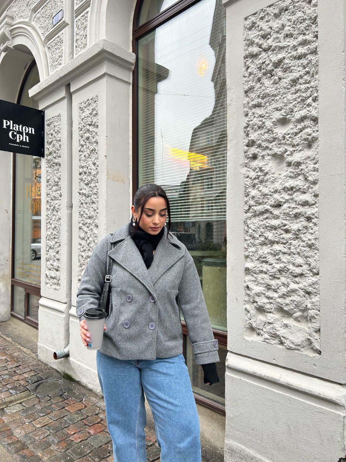 Frau in grauem Mantel mit Kaffeebecher vor Platon Cph Geschäft, modisch, urbaner Stil.