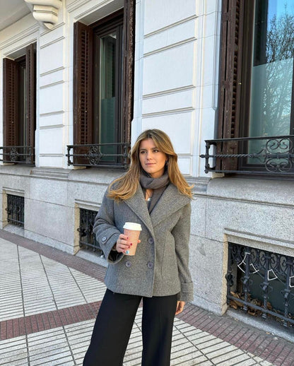Frau in grauem Wollmantel mit Kaffeebecher, steht vor einem Gebäude, Herbstmode.