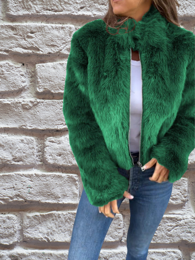Frau in grüner Kunstpelzjacke, weiße Bluse, blaue Jeans, vor Ziegelwand.