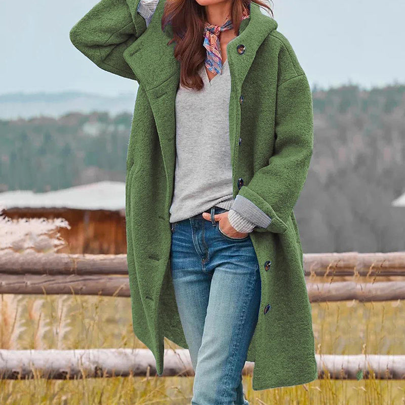 Frau in grünem Wollmantel, grauem Pullover und Jeans vor ländlicher Kulisse.
