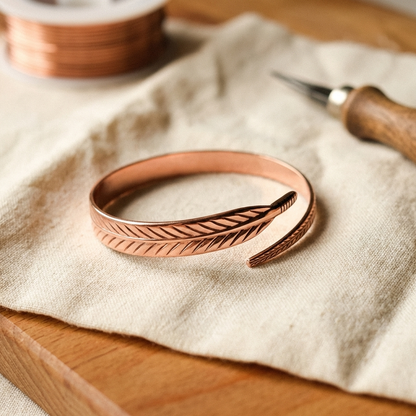 Offenes Kupferarmband mit eingraviertem Federmuster liegt auf einem Leinentuch, daneben eine Drahtspule und ein Gravierwerkzeug mit Holzgriff. 1