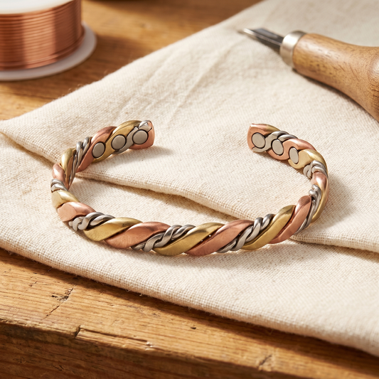 Offenes, dreisträngig gedrehtes Armband aus Rosékupfer, Messing und silberfarbenem Metall auf Leinentuch, daneben 1
