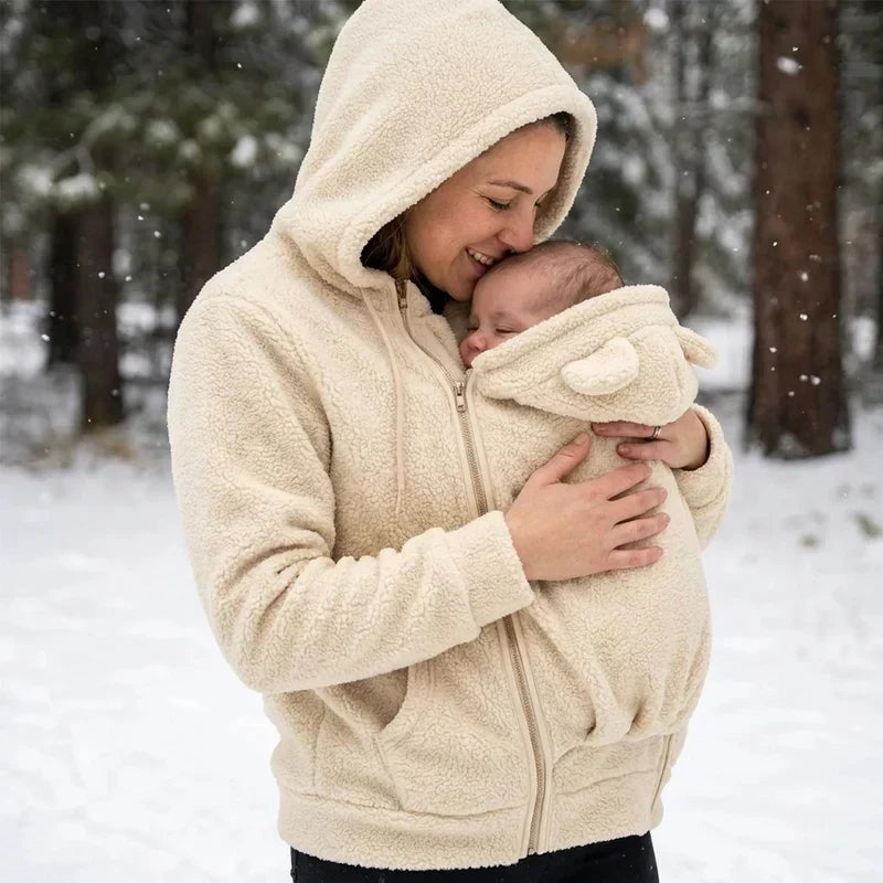 Lächelnde Frau in cremefarbenem Teddyfleece-Pullover mit Kapuze küsst ihr Baby im verschneiten Wald.