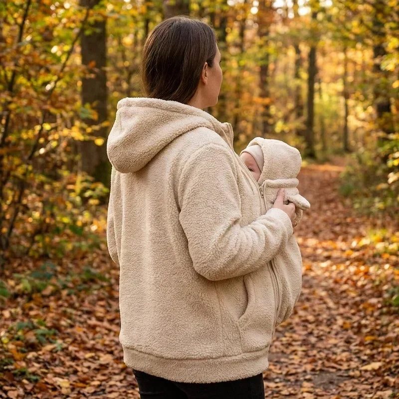 Seitenansicht einer Frau im cremefarbenen Fleece-Kapuzenpullover, die im Wald ein Baby vorn im Pullover trägt.