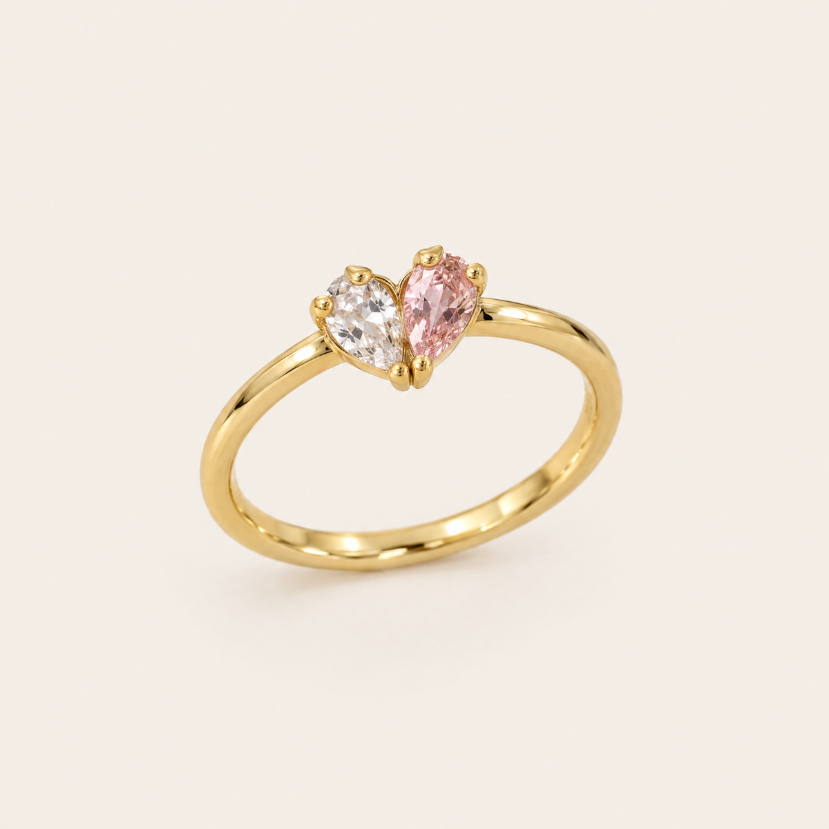 Goldfarbener Ring von schräg oben, zwei tropfenförmige Steine in Klar und Rosa bilden gemeinsam ein Herz auf dem schmalen Ringband. 1
