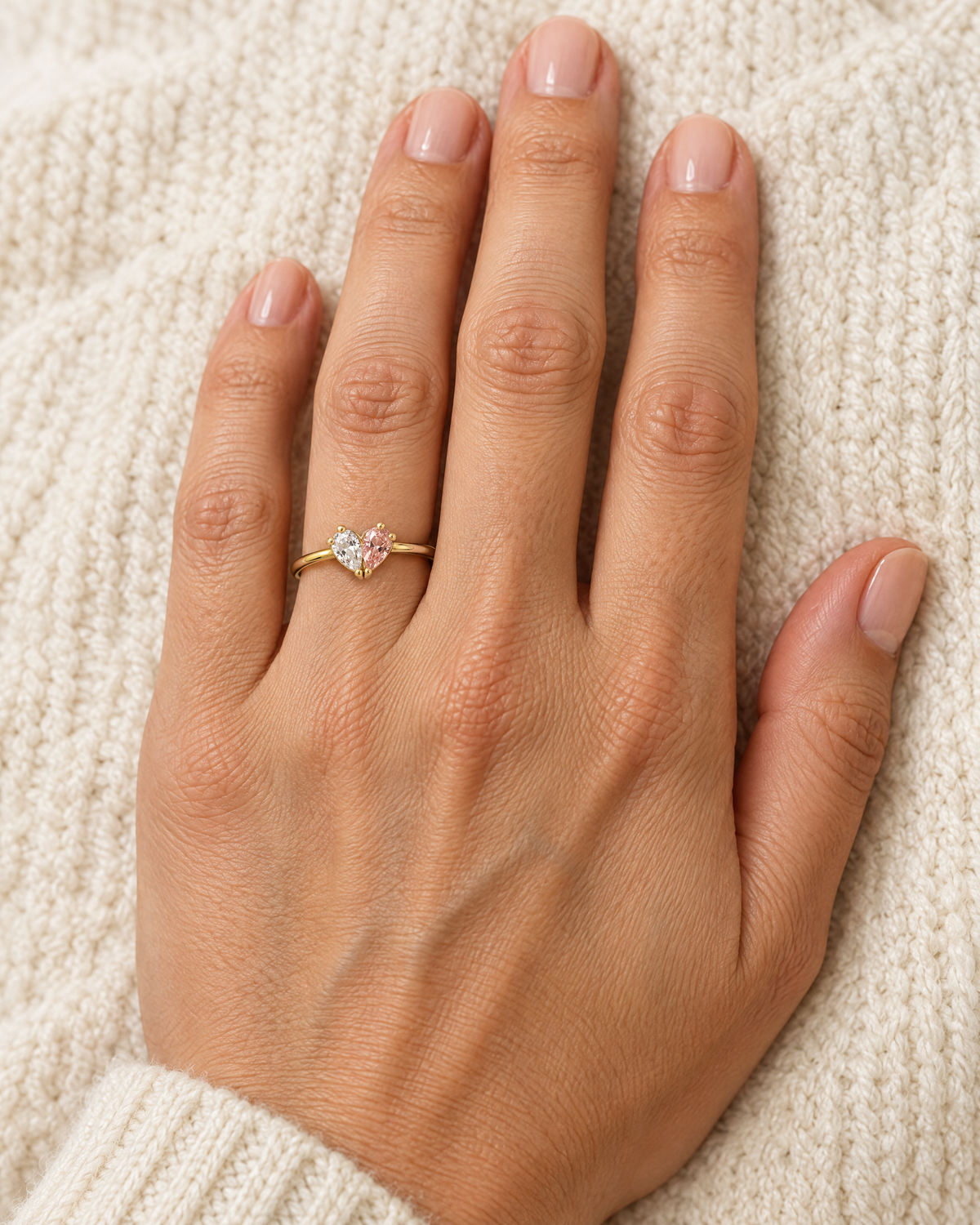 Der Ring mit klarem und rosa Stein am Finger einer Hand, die auf einem cremefarbenen Strickstoff liegt. 2