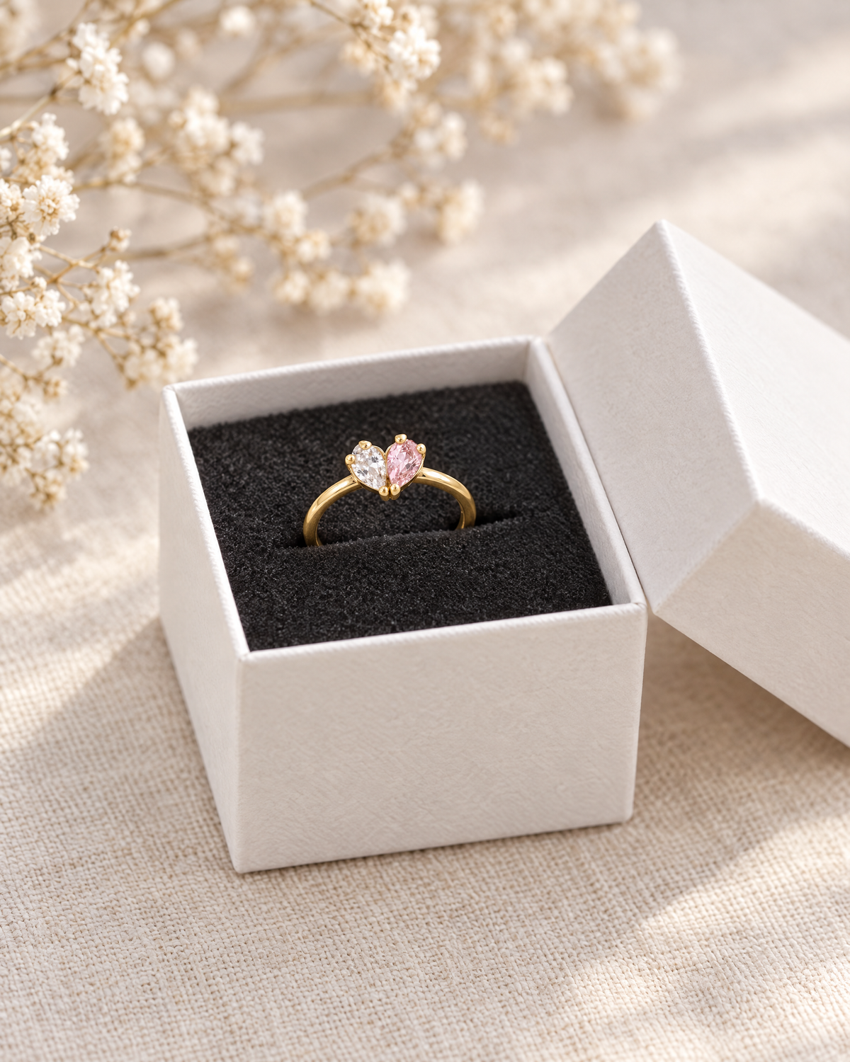Der goldfarbene Herzring in einer weißen Schmuckschachtel mit schwarzem Einsatz, daneben getrocknete weiße Blüten als Dekoration. 3