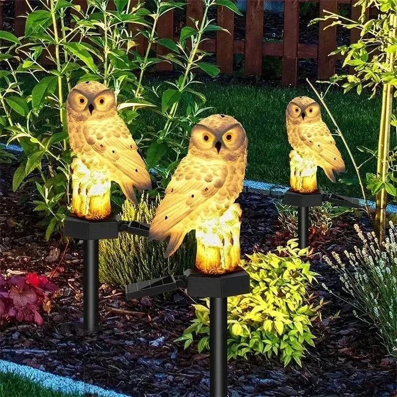 Drei braun-beige Eulen-Gartenlichter mit leuchtendem Baumstumpf-Sockel im Beet.