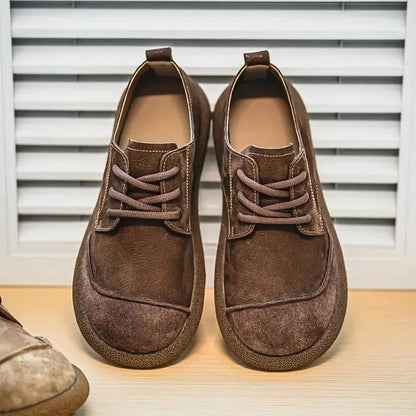Paar dunkelbraune Schnürschuhe von vorn auf einem Holzbrett vor einer Lamellenwand, links ragt ein hellerer Schuh ins Bild. 6