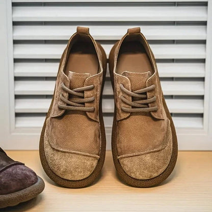 Paar hellbraune Schnürschuhe von vorn auf einem Holzbrett vor einer Lamellenwand, links ein dunklerer Schuh am Bildrand. 7