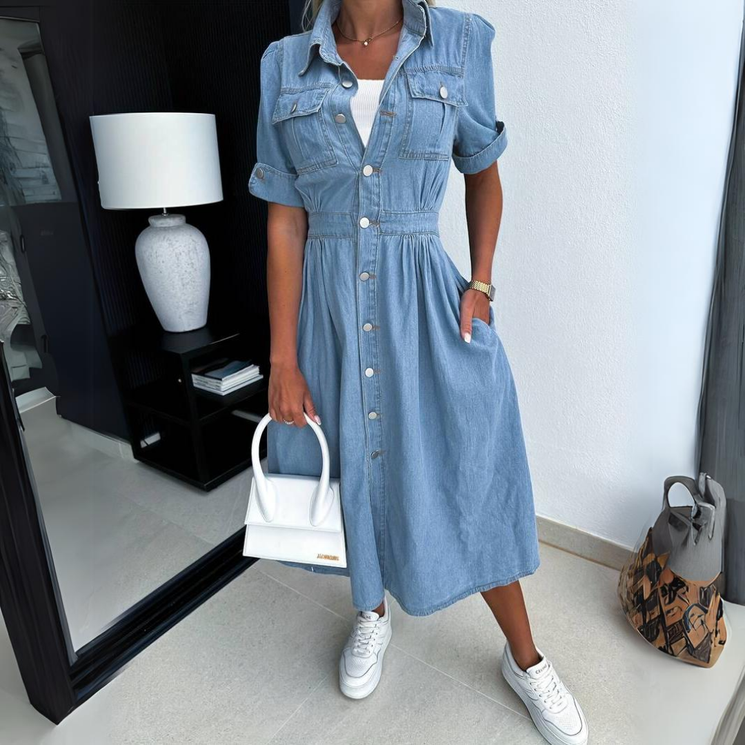 Frau in blauem Jeanskleid mit weißen Sneakers und weißer Handtasche in modernem Raum.