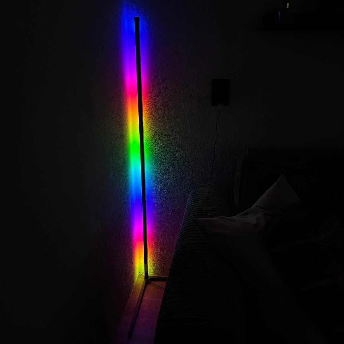 Eckstehlampe im abgedunkelten Raum mit buntem RGB-Farbverlauf an der Wand, rechts ein Sofa und ein Netzstecker mit Kabel. 6