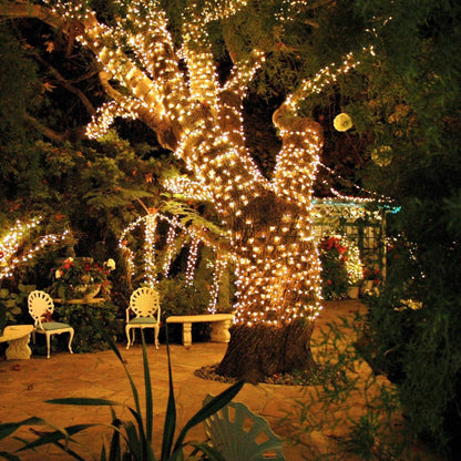 Beleuchteter Baum im romantischen Garten mit Lichterketten und weißen Gartenstühlen.