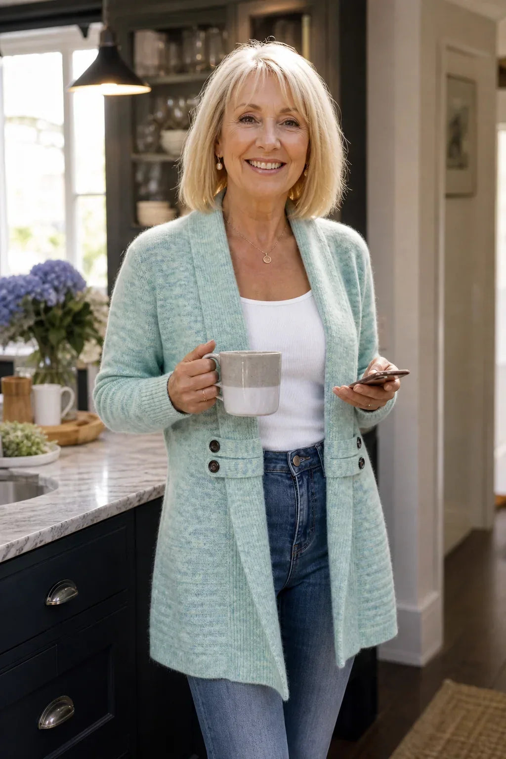 Lächelnde Frau in der Küche trägt die mintgrüne Strickjacke mit Tasse und Smartphone in der Hand 4