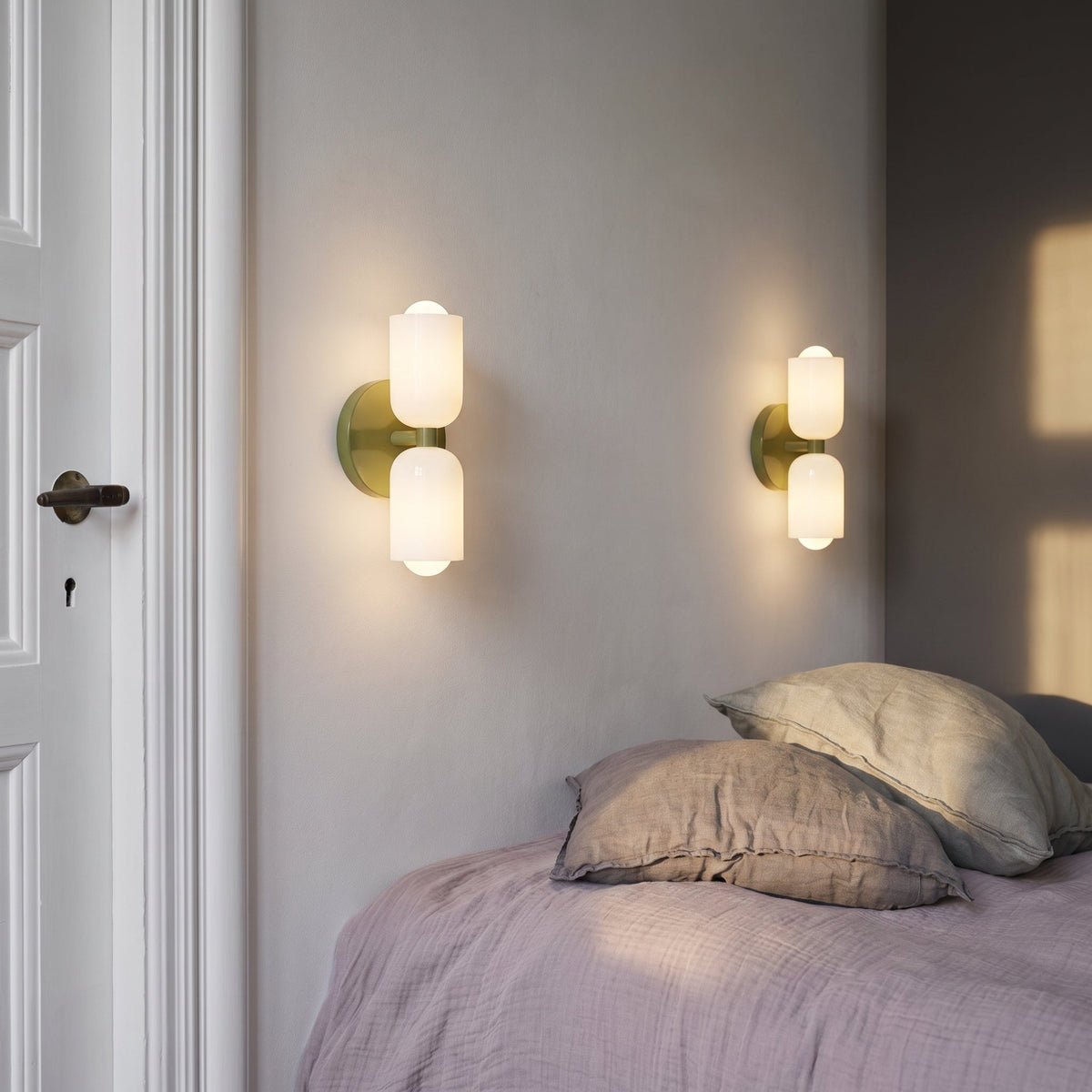 Zwei Wandleuchten mit olivgrüner Metallplatte und weißen Glasschirmen über einem Bett mit zerknitterter Leinenbettwäsche in Rosa.