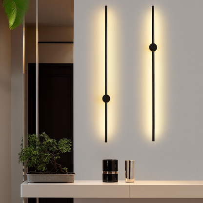 Zwei schwarze Stab-Wandleuchten mit warmem Lichtschein an grauer Wand über einem hellen Sideboard mit Bonsai und Vasen.