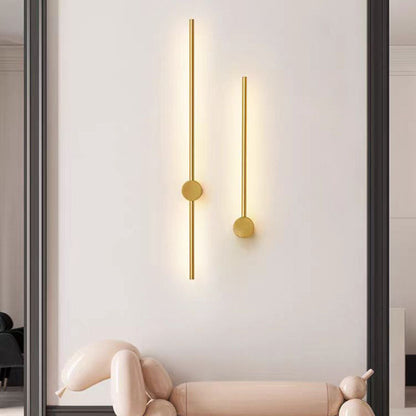 Zwei goldfarbene Stab-Wandleuchten unterschiedlicher Länge mit runder Wandplatte, warm leuchtend, an einer hellen Wand über einem rosa Ballontier.