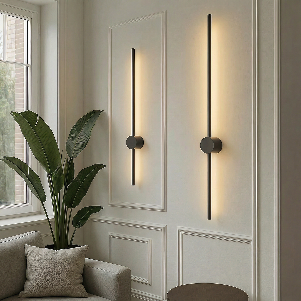 Zwei schwarze, unterschiedlich lange Stab-Wandleuchten mit warmem Licht an einer weißen Stuckwand neben Zimmerpflanze und Sofa.