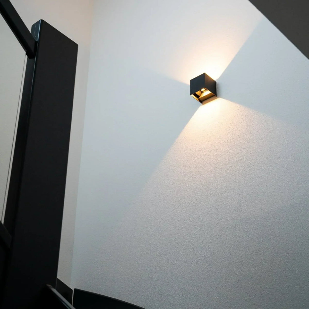 Schwarze Wandleuchte an weißer Treppenhauswand, schräg von unten fotografiert, mit Lichtkegeln nach oben und unten 8