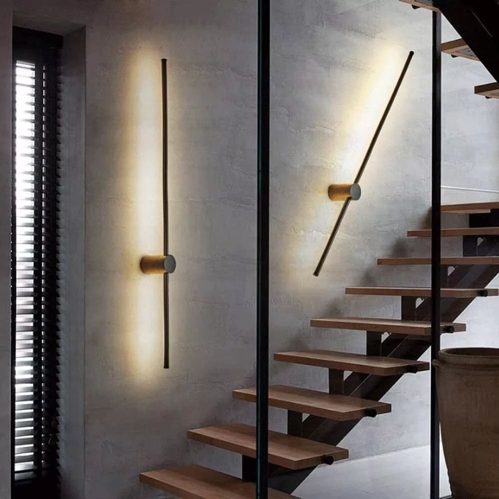 Eine senkrechte und eine schräg gestellte Stableuchte mit goldfarbenem Zylinder an einer Betonwand neben einer Treppe mit Holzstufen. 4