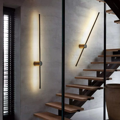 Eine senkrechte und eine schräg gestellte Stableuchte mit goldfarbenem Zylinder an einer Betonwand neben einer Treppe mit Holzstufen. 4