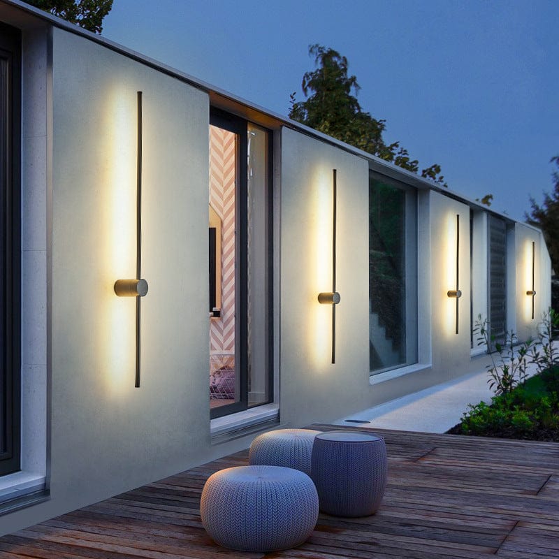 Mehrere schwarze lineare LED-Wandleuchten leuchten abends warmweiß an einer modernen Hauswand oberhalb einer Holzterrasse mit zwei gestrickten Sitzpoufs. 1