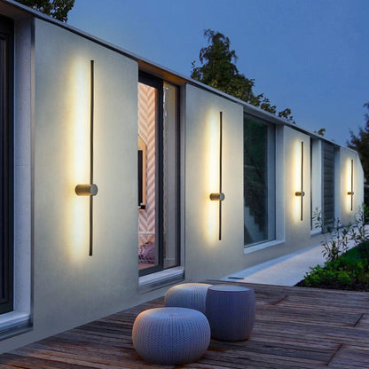 Mehrere schwarze lineare LED-Wandleuchten leuchten abends warmweiß an einer modernen Hauswand oberhalb einer Holzterrasse mit zwei gestrickten Sitzpoufs. 1